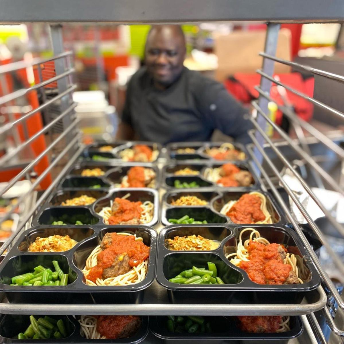 A cart is stacked full of trays of school meals - spaghetti and meatballs, green beans, and apple sauce. A chef smiles in the background. A cart is stacked full of trays of school meals - spaghetti and meatballs, green beans, and apple sauce. A chef smiles in the background.