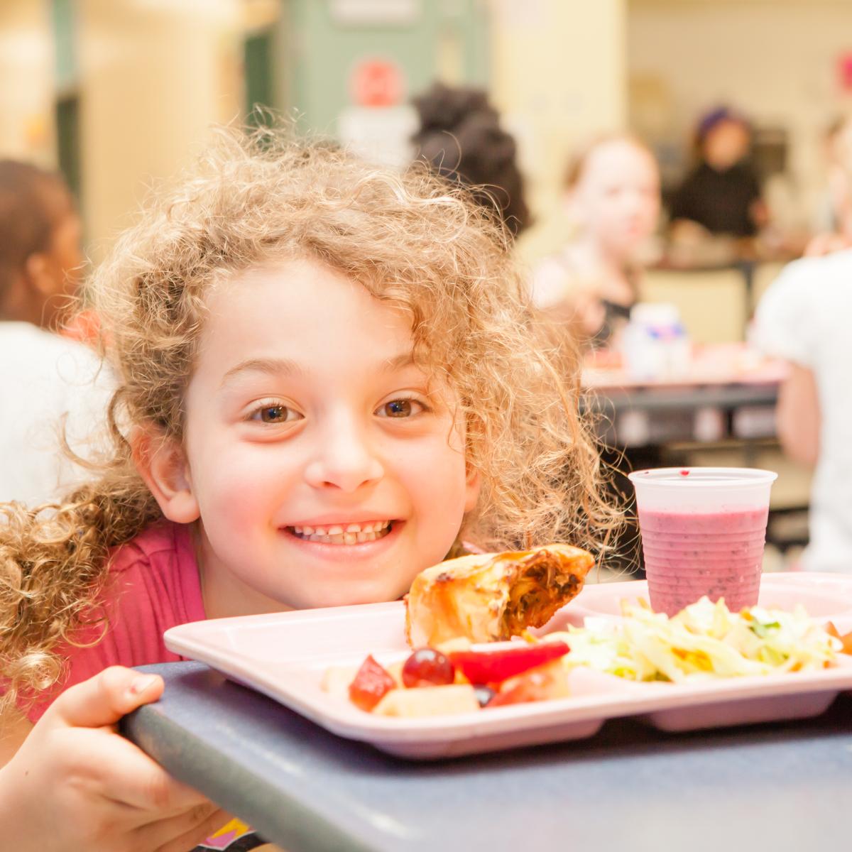 A child smiles, a lunch tray sitting on the cafeteria table in front of them. A child smiles, a lunch tray sitting on the cafeteria table in front of them.