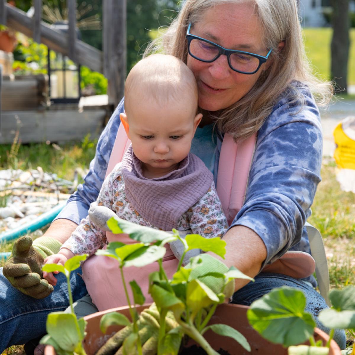 A woman sits with a baby in a carrier attached to her body, they look at a squash plant A woman sits with a baby in a carrier attached to her body, they look at a squash plant