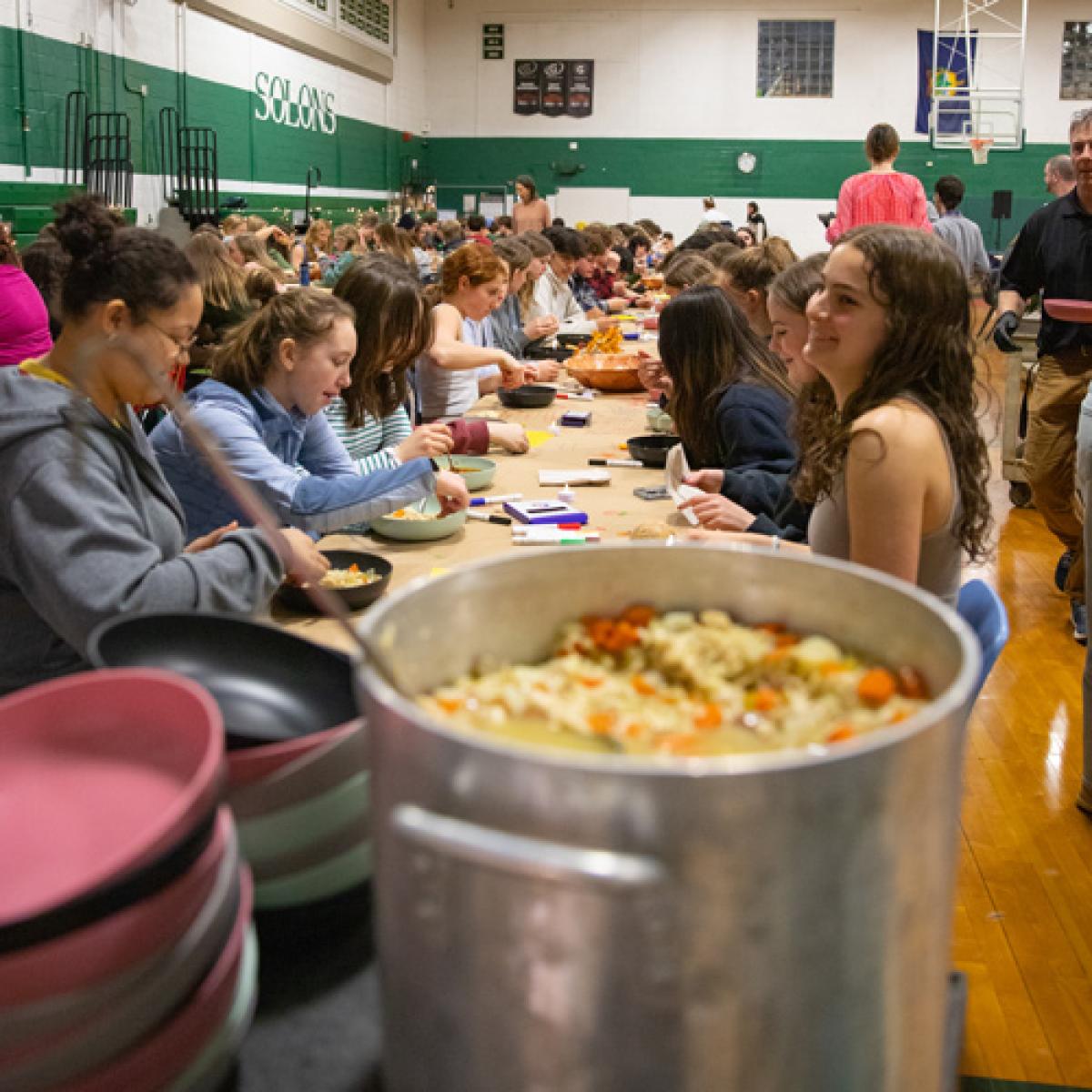 A large group of students dining together at long tables in a gymnasium. Several large pots of soup are placed on the tables. The setting suggests a communal or celebratory event. A large group of students dining together at long tables in a gymnasium. Several large pots of soup are placed on the tables. The setting suggests a communal or celebratory event.