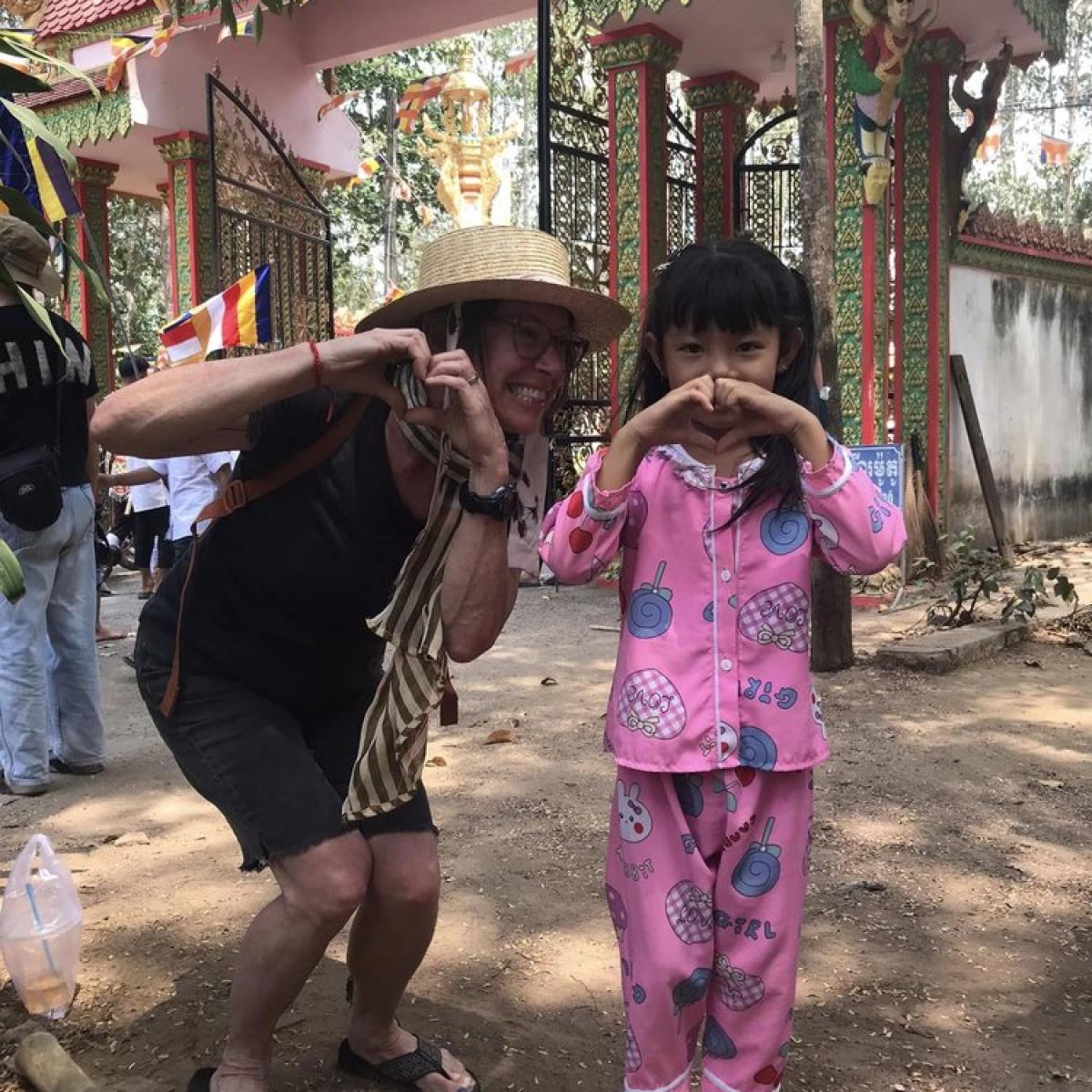 A person in a straw hat and shorts making a peace sign next to a child wearing pink pajamas and forming a heart shape with their hands, both smiling, standing in front of a temple setting with intricate structures and trees. A person in a straw hat and shorts making a peace sign next to a child wearing pink pajamas and forming a heart shape with their hands, both smiling, standing in front of a temple setting with intricate structures and trees.