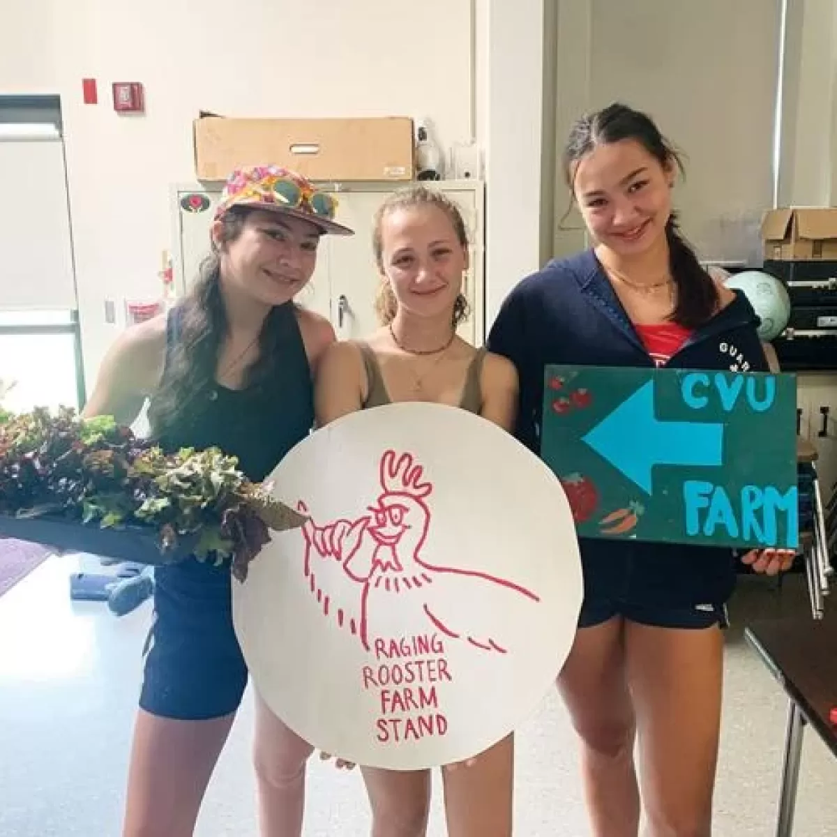 Three students holding items related to a farm stand. The person on the left is holding a large bunch of leafy greens, and the person in the middle holds a round sign with the image of a rooster and the text "RAGING ROOSTER FARM STAND." The individual on the right holds a blue arrow sign with "CVU FARM" written on it. Three students holding items related to a farm stand. The person on the left is holding a large bunch of leafy greens, and the person in the middle holds a round sign with the image of a rooster and the text "RAGING ROOSTER FARM STAND." The individual on the right holds a blue arrow sign with "CVU FARM" written on it.