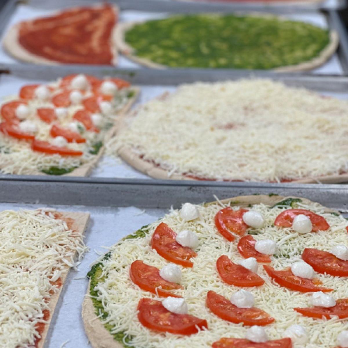 Several unbaked pizzas on trays, each topped with different ingredients such as tomato sauce, pesto, shredded cheese, sliced tomatoes, and dollops of cheese. Several unbaked pizzas on trays, each topped with different ingredients such as tomato sauce, pesto, shredded cheese, sliced tomatoes, and dollops of cheese.