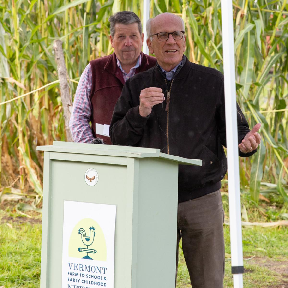 Two individuals standing at a podium with the "Vermont Farm to School and Early Childhood" logo, located in an outdoor setting with cornstalks in the background. Two individuals standing at a podium with the "Vermont Farm to School and Early Childhood" logo, located in an outdoor setting with cornstalks in the background.