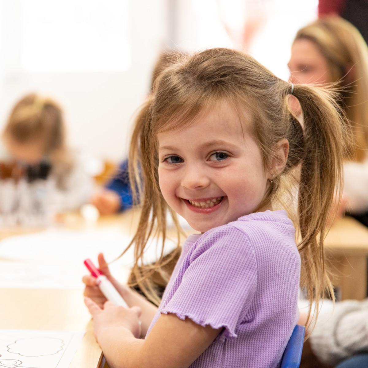 A smiling child with a ponytail sits at a table holding a marker, in a classroom setting. A smiling child with a ponytail sits at a table holding a marker, in a classroom setting.