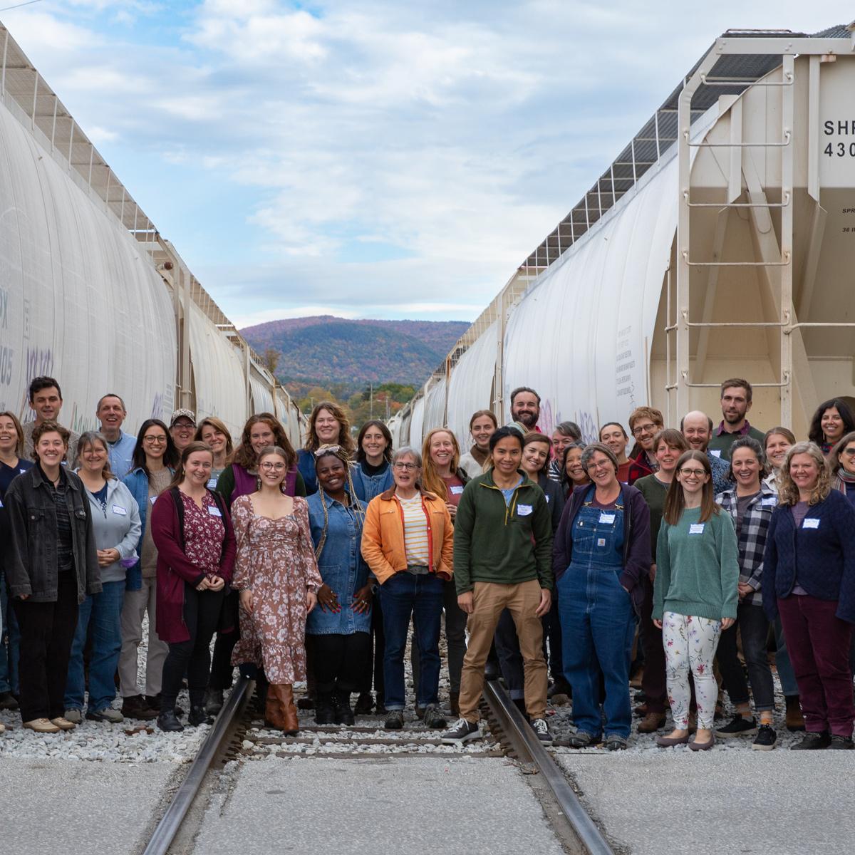 Group of people standing between two rows of white train cars on railroad tracks, with a scenic mountain backdrop. Group of people standing between two rows of white train cars on railroad tracks, with a scenic mountain backdrop.