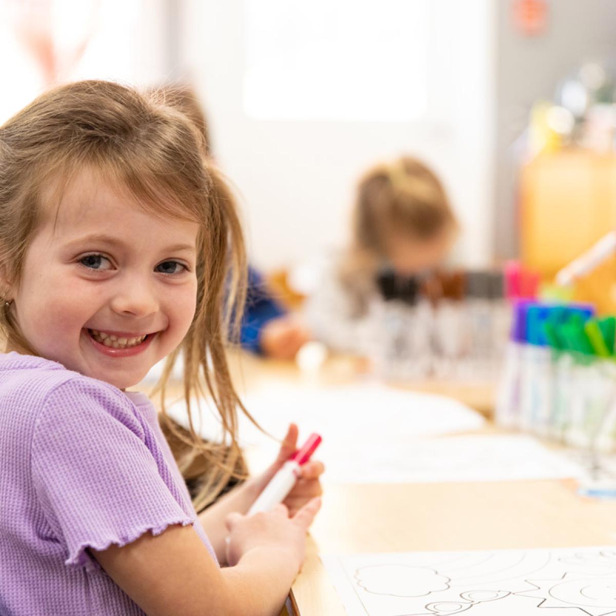 Child with a smile, holding a crayon at a classroom table, with colorful art supplies in the background and other children engaged in activities.