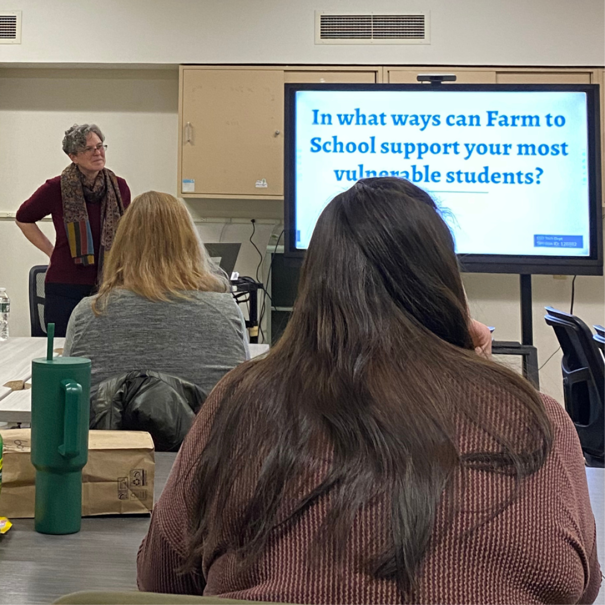 Presenter standing next to a screen displaying a presentation titled 'In what ways can Farm to School support your most vulnerable students?' with attendees seated and listening in a classroom setting. Presenter standing next to a screen displaying a presentation titled 'In what ways can Farm to School support your most vulnerable students?' with attendees seated and listening in a classroom setting.