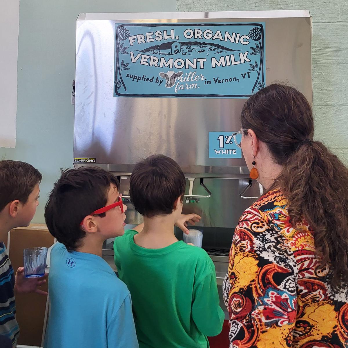 Three children and one adult at a milk dispensing station with a sign reading "Fresh, Organic Vermont Milk supplied by Miller Farm in Vernon, VT." Three children and one adult at a milk dispensing station with a sign reading "Fresh, Organic Vermont Milk supplied by Miller Farm in Vernon, VT."