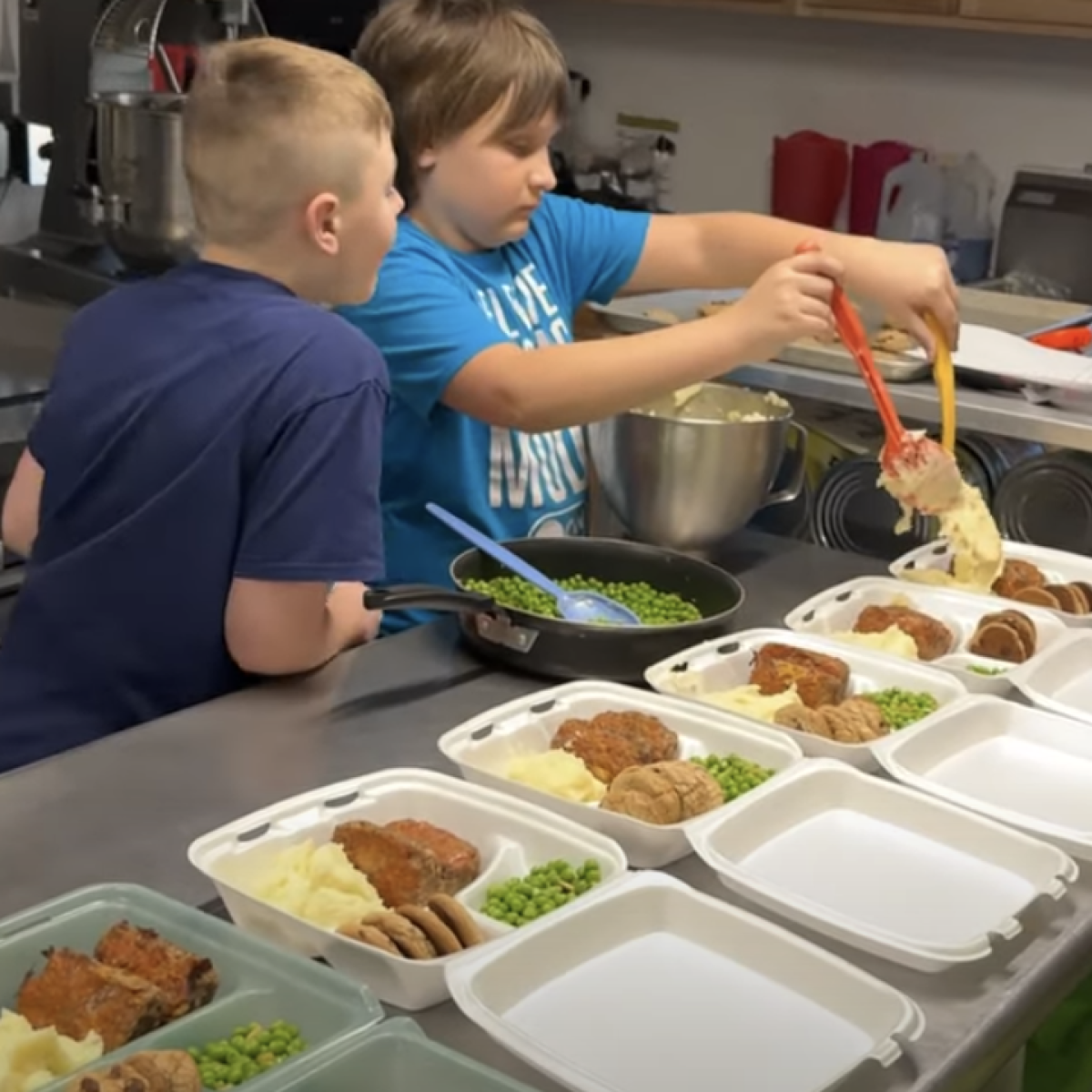 Two children preparing meals in a kitchen. Multiple dishes with food are arranged on the counter, including meatloaf, peas, and mashed potatoes. Two children preparing meals in a kitchen. Multiple dishes with food are arranged on the counter, including meatloaf, peas, and mashed potatoes.