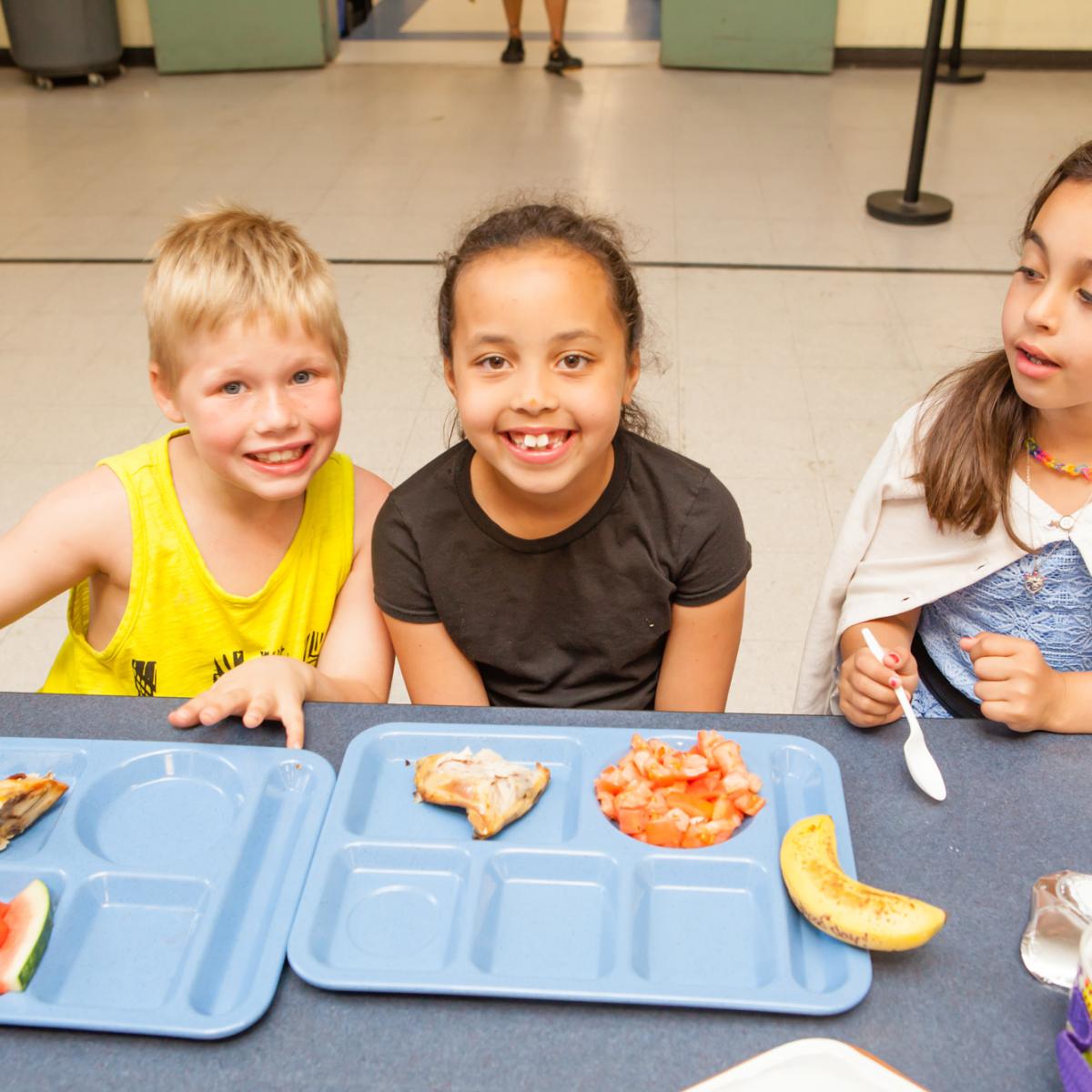 Children smiling at a lunch table with trays of food in a school cafeteria. Children smiling at a lunch table with trays of food in a school cafeteria.
