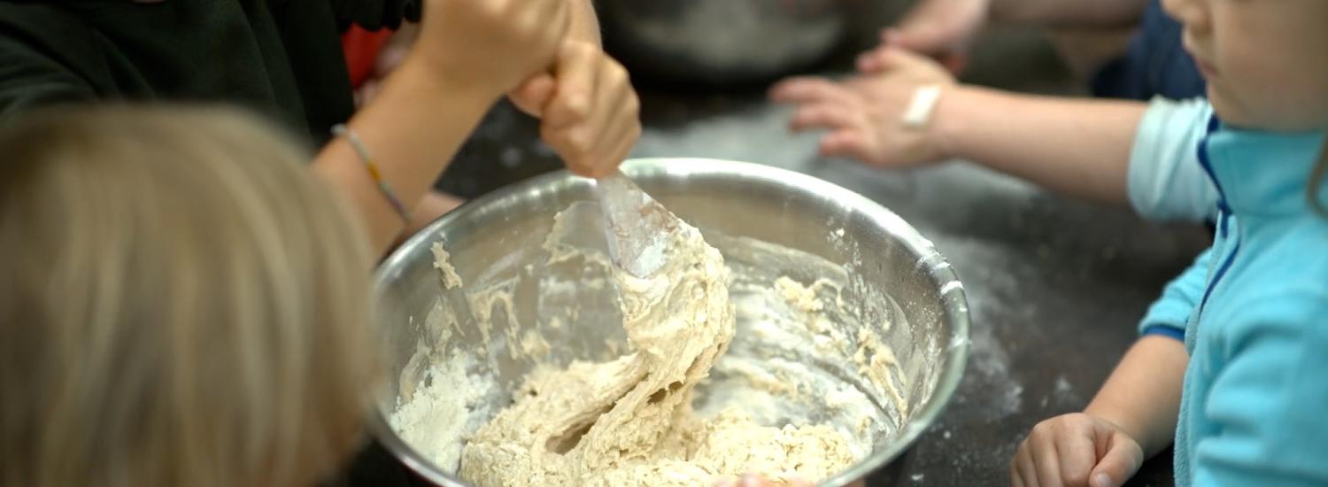 Seen from above, students mix bread dough in a large stainless steel bowl with a wooden spoon.